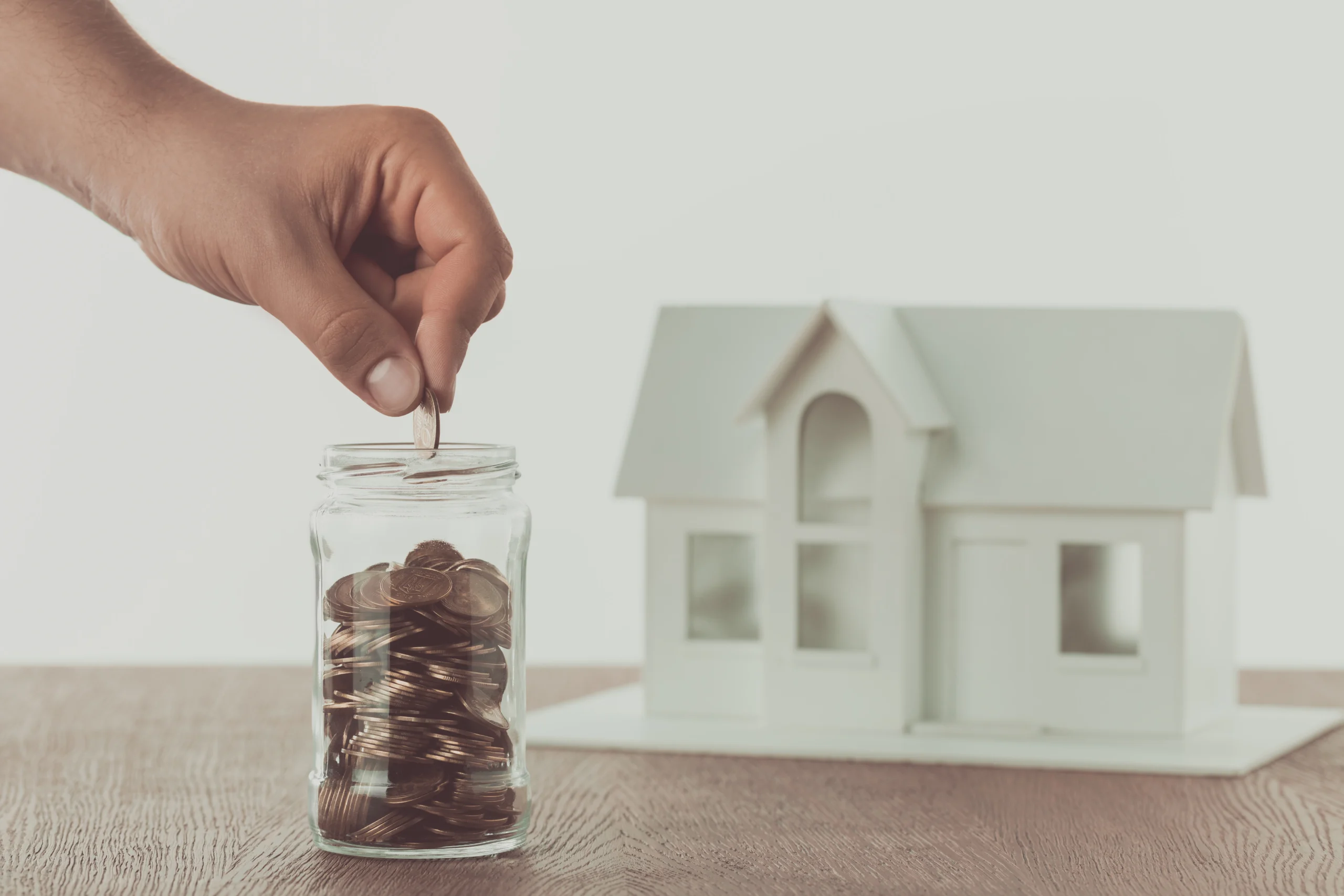 cropped image of man putting coin into glass jar with small house on table, saving concept